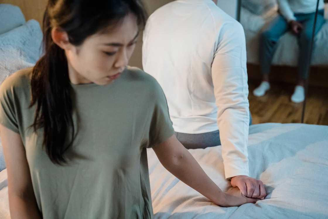 A woman and a man sitting back-to-back on a bed, their hands touching despite visible emotional distance.