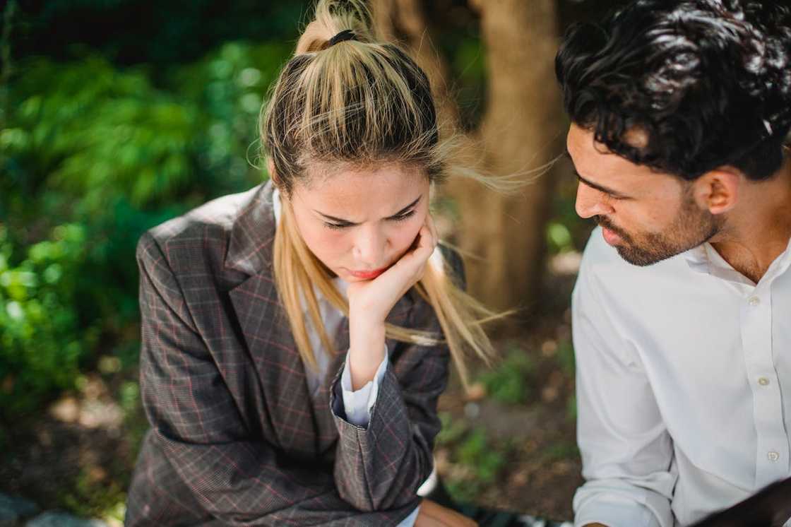 A woman in a blazer looks distressed while a man speaks to her outdoors.