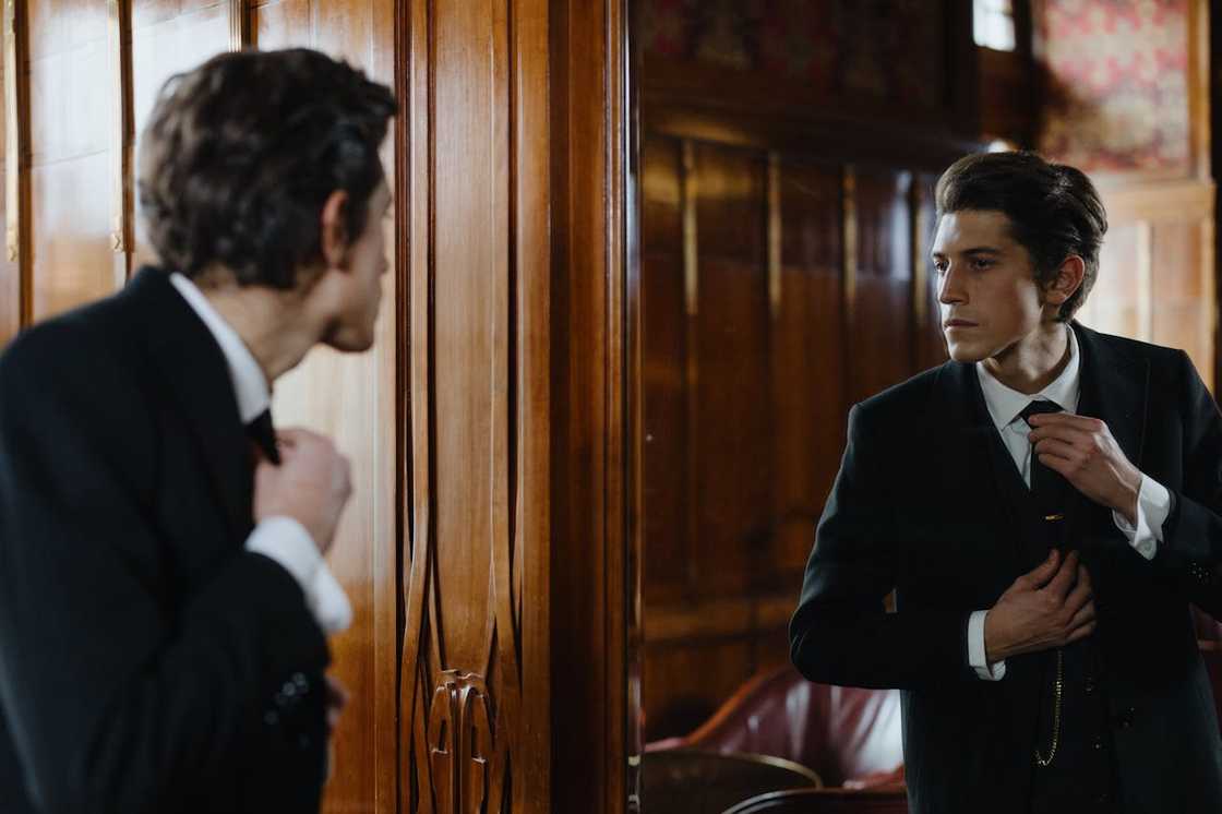A man in a black suit adjusts his tie while looking at his reflection in a mirror.