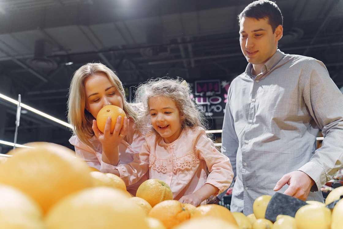 A woman smells an orange while a smiling child reaches for fruit, with a man standing beside them.