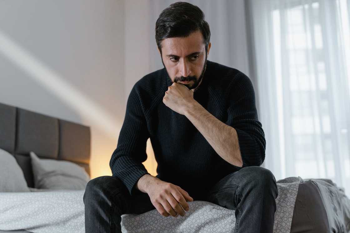 A man sits on the edge of a bed, resting his chin on his hand and looking down thoughtfully. A man sits on the edge of a bed, resting his chin on his hand and looking down thoughtfully.
