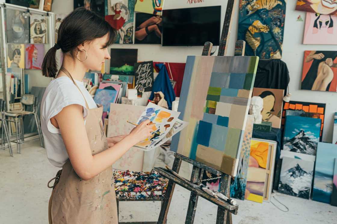 A woman in a paint-splattered apron works on a geometric abstract painting in a studio.