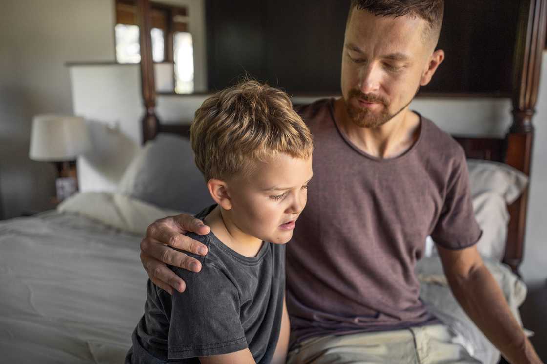 Father laying arm around his sad son on bed at home Father laying arm around his sad son on bed at home