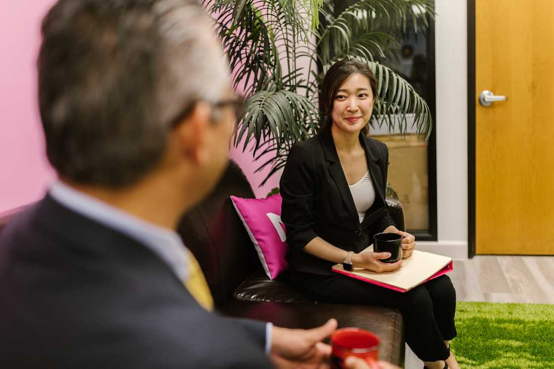 A woman holding a notebook and cup, listening during a conversation in an office. A woman holding a notebook and cup, listening during a conversation in an office.