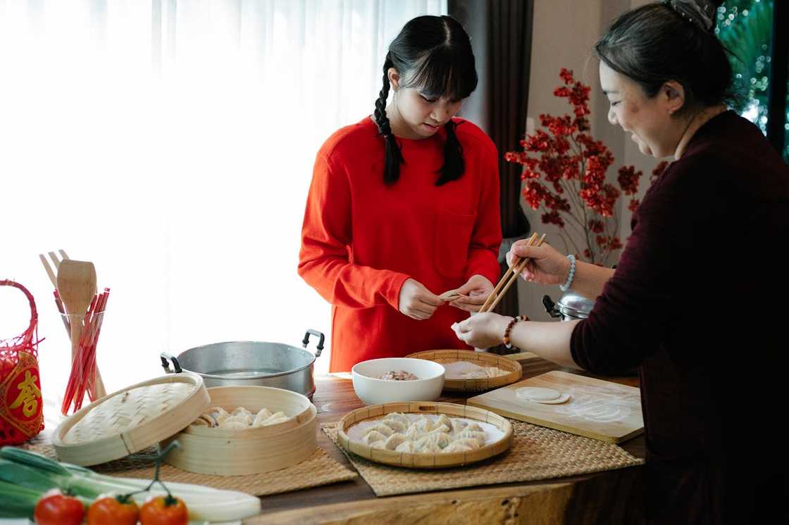 Two people prepare dumplings together at a kitchen table filled with bamboo steamers.