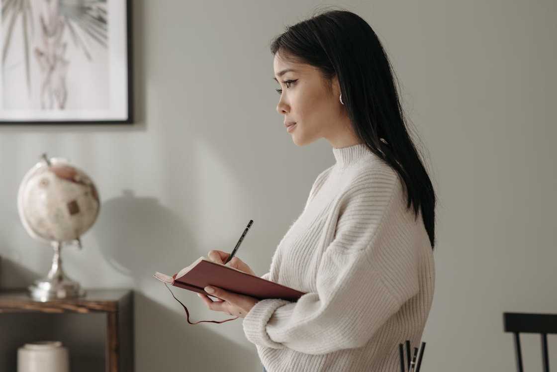A woman stands indoors writing in a notebook while looking ahead thoughtfully.