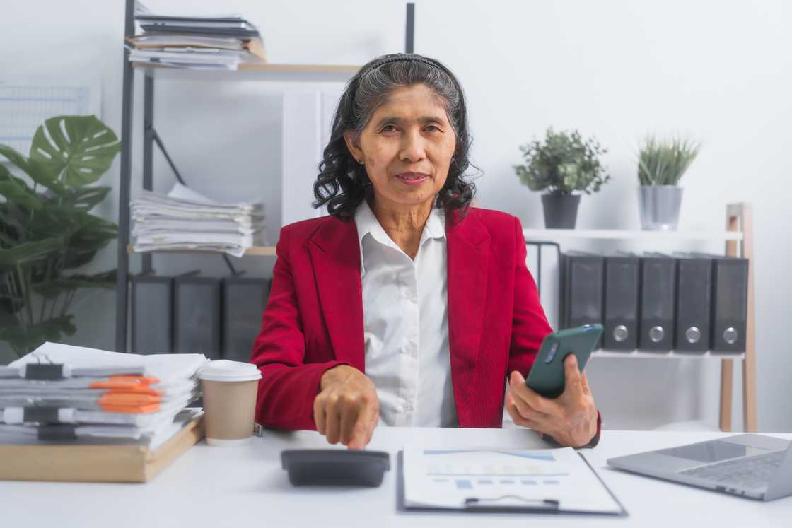 Smiling elderly woman seated at a desk in an archive workspace.