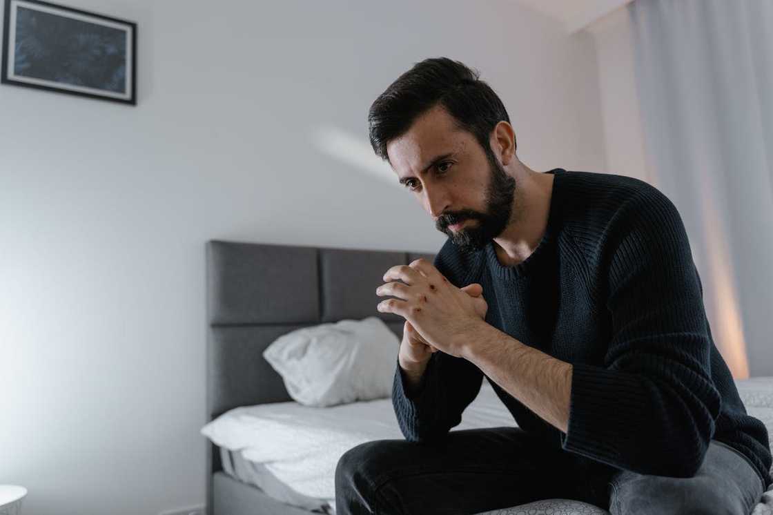 A man sits on the edge of a bed with his hands clasped, looking down thoughtfully in a quiet bedroom. A man sits on the edge of a bed with his hands clasped, looking down thoughtfully in a quiet bedroom.