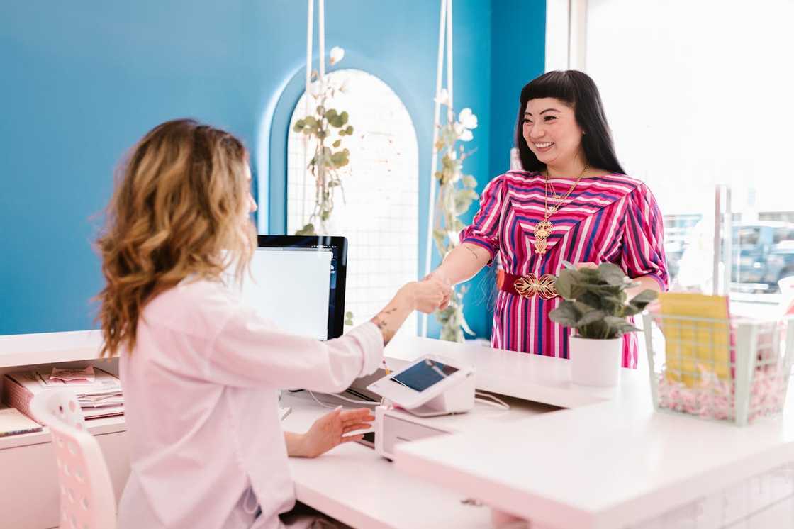 A smiling woman in a colourful striped dress shakes hands with another woman across a white desk.