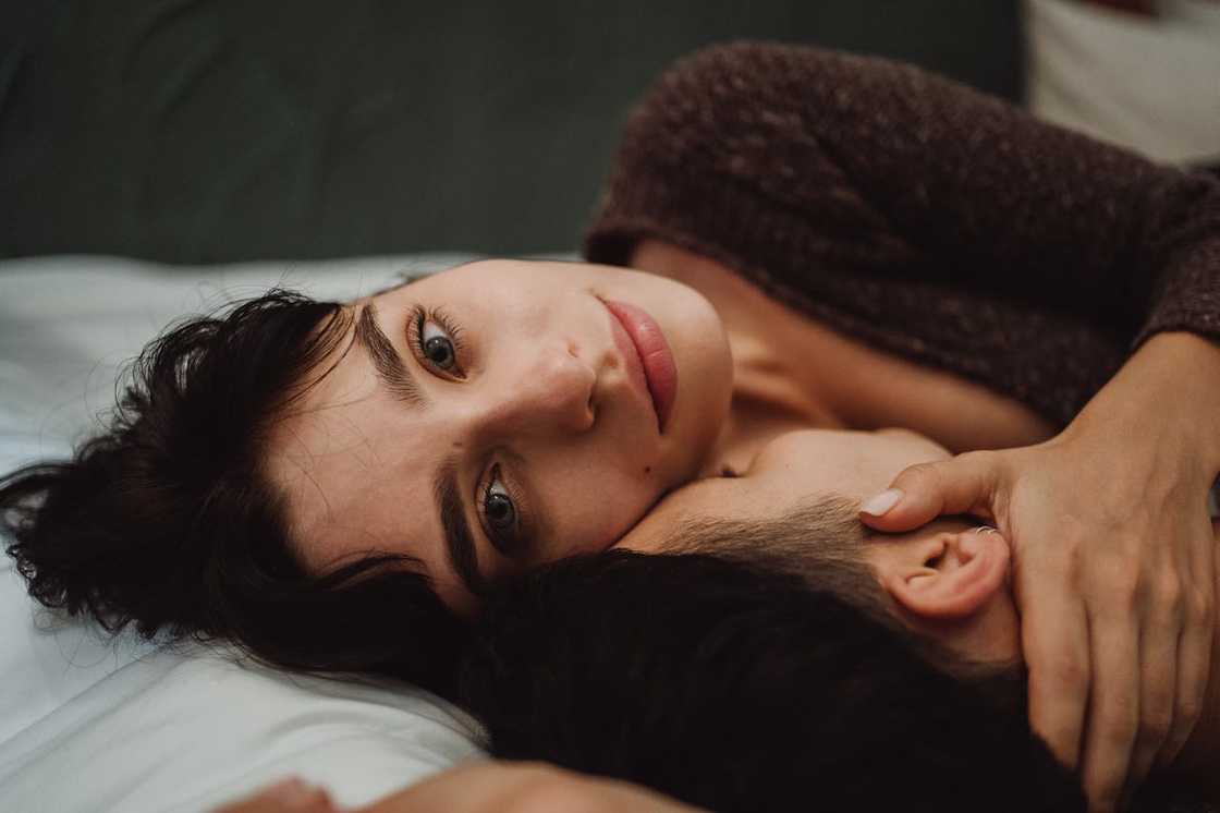 A woman lies on a bed holding a man close, gazing ahead with a calm, expression.