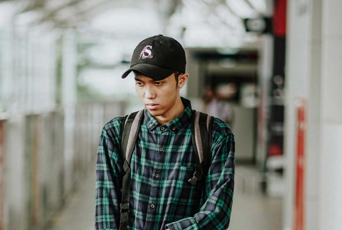 A young person in a plaid shirt and black cap stands in a covered walkway.