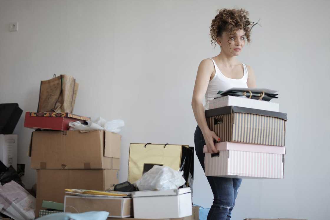 A woman carries a stack of boxes while standing in a room filled with packed belongings.