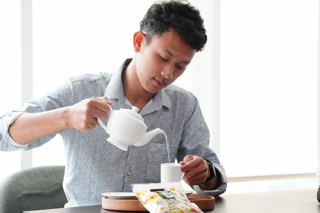 A man pours hot water from a teapot into a cup while preparing a drink at a table. A man pours hot water from a teapot into a cup while preparing a drink at a table.