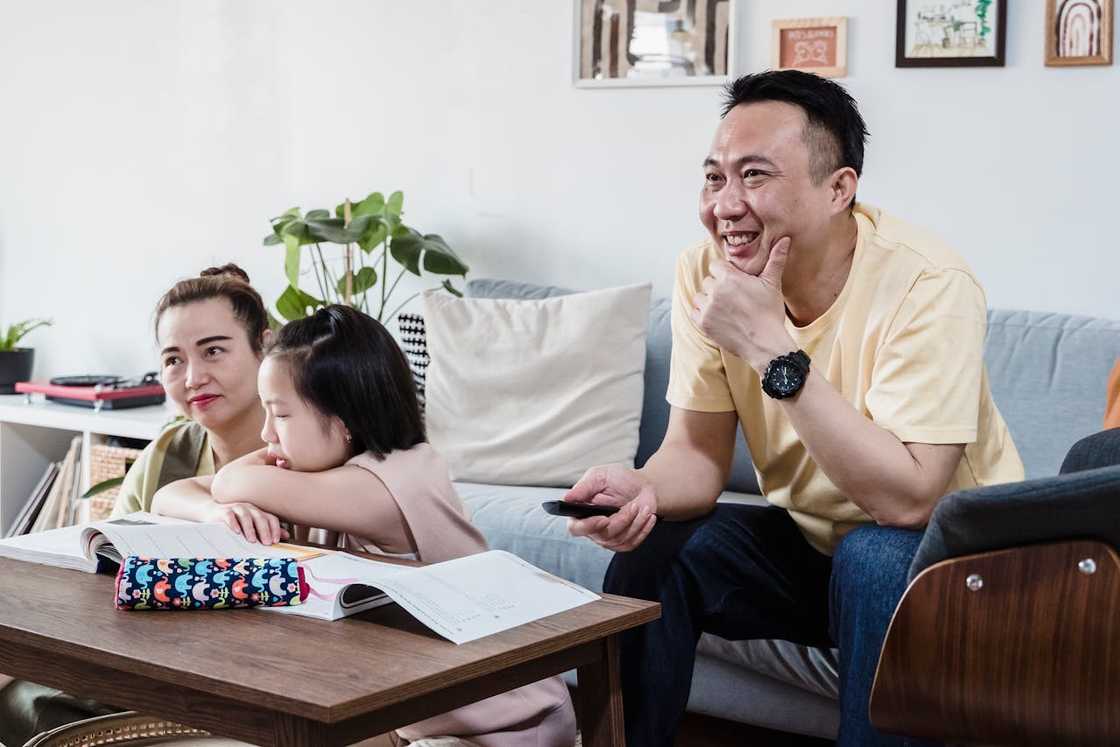 A young girl studies at a table with open books while two adults sit nearby. A young girl studies at a table with open books while two adults sit nearby.