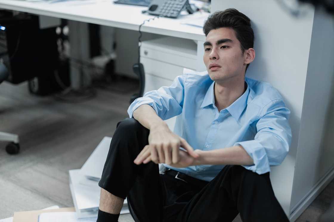 A stressed office worker sitting on the floor beside a desk, looking exhausted.