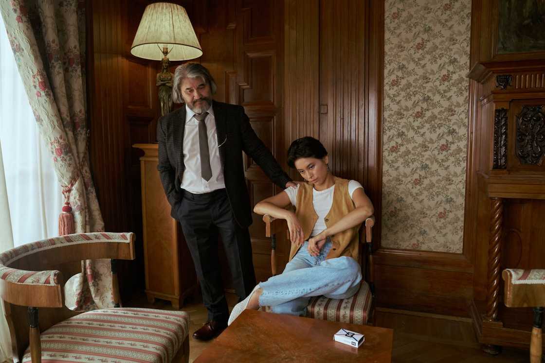 An older man in a suit stands behind a seated young man in a wood-panelled room.