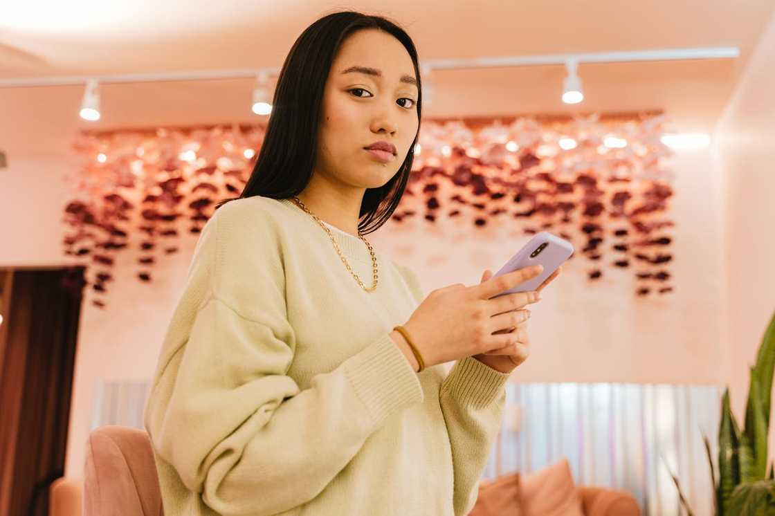 A woman stands indoors holding a smartphone, looking serious. A woman stands indoors holding a smartphone, looking serious.