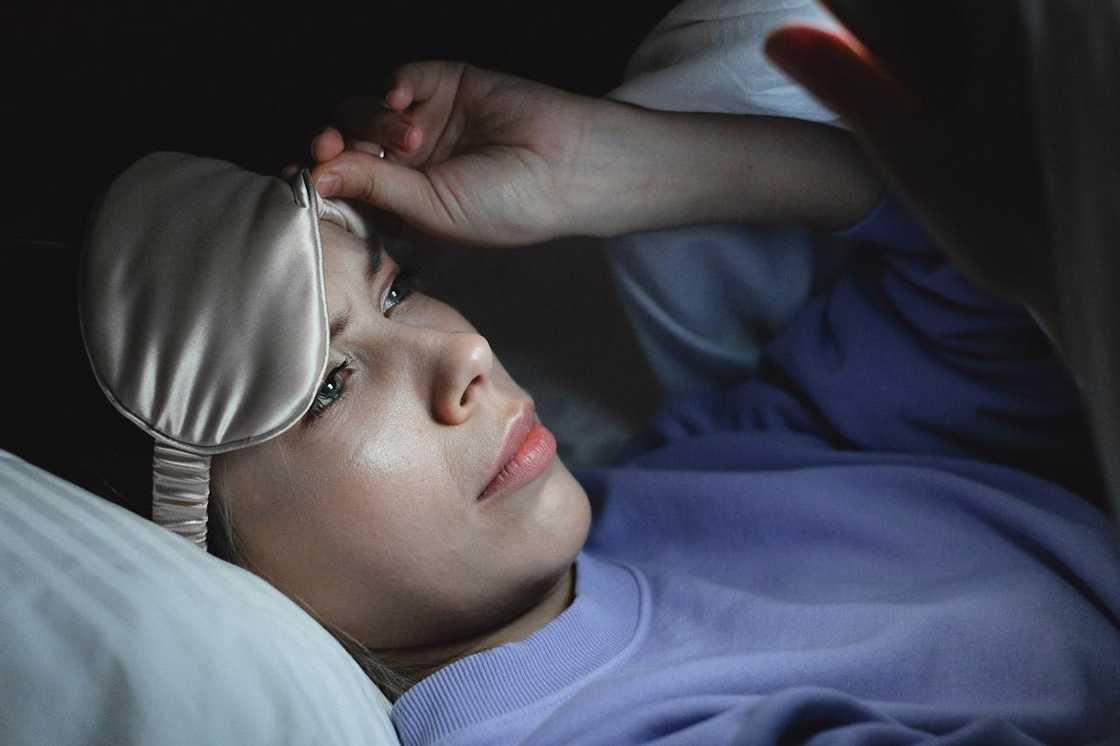 A woman lies awake in bed, wearing a sleep mask and staring at her phone. A woman lies awake in bed, wearing a sleep mask and staring at her phone.