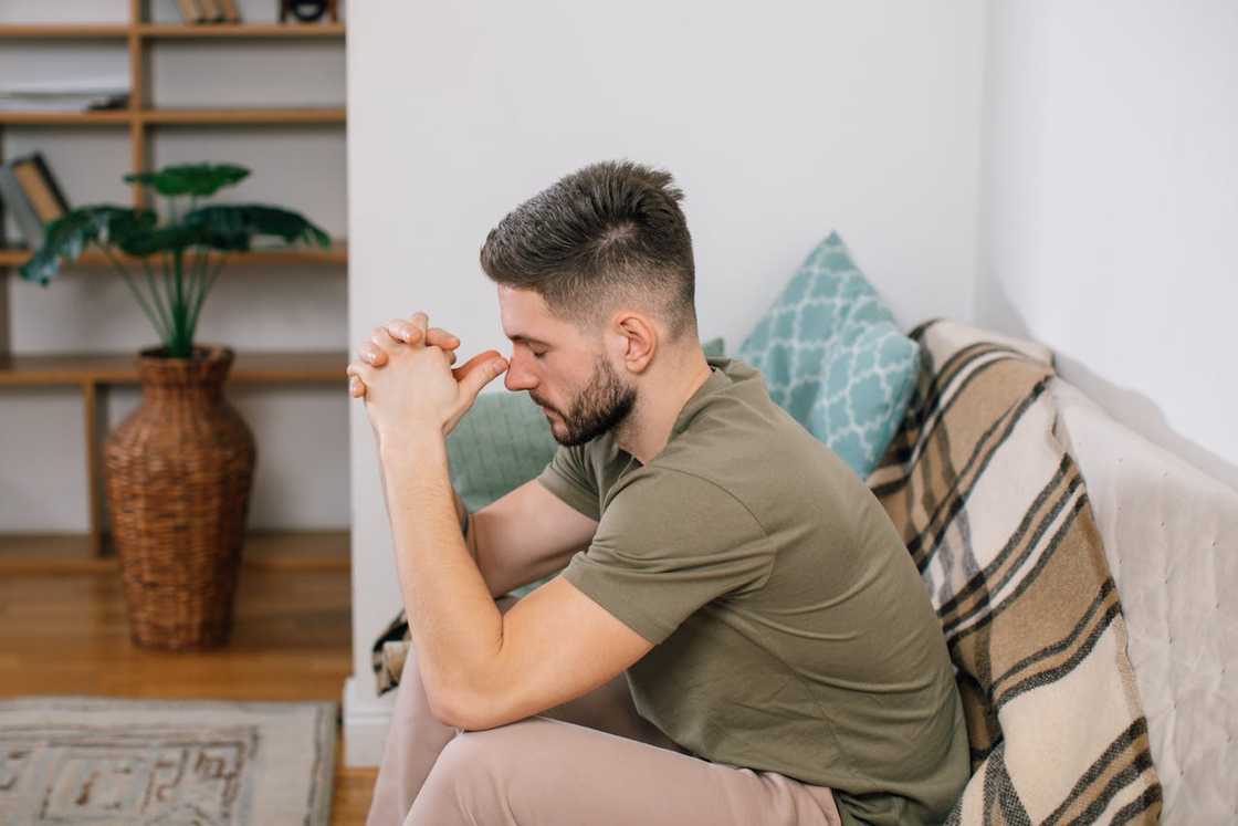A man sits on a couch with his hands clasped, looking down in deep thought. A man sits on a couch with his hands clasped, looking down in deep thought.