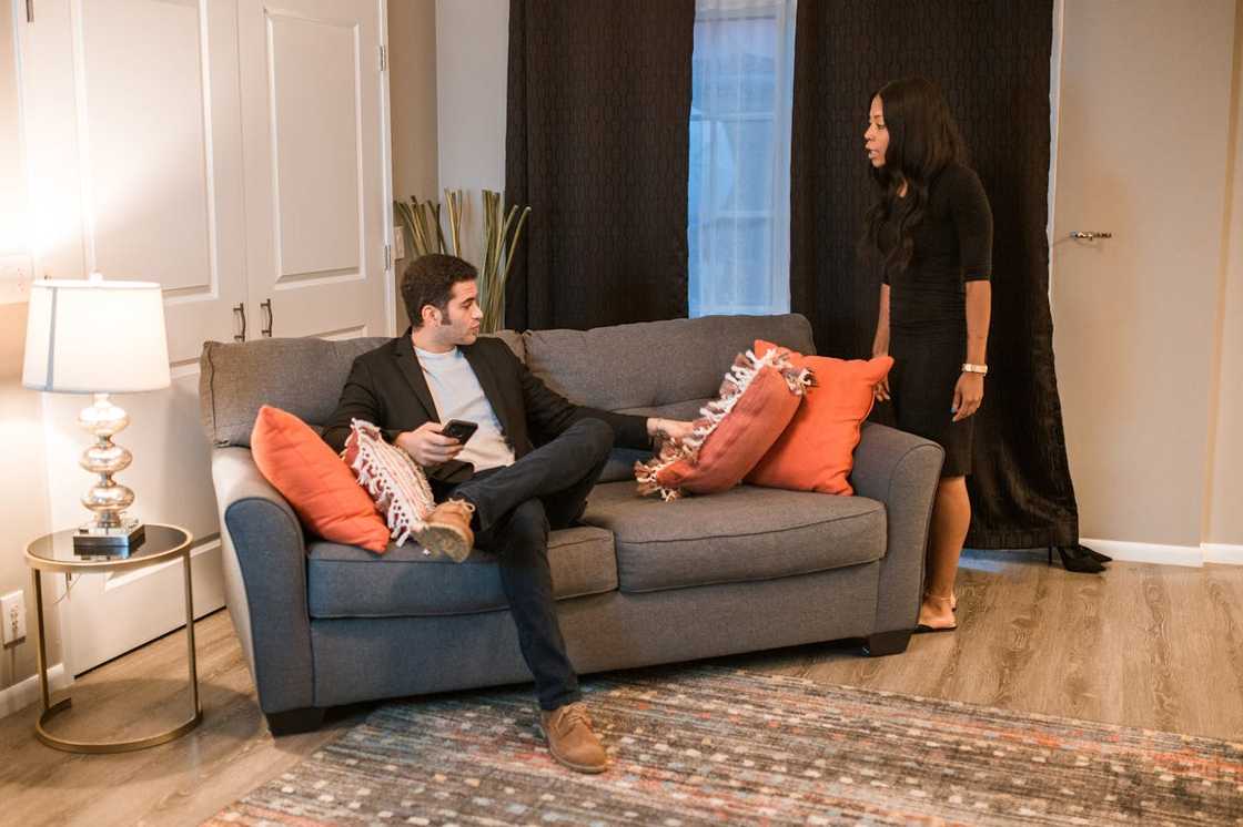 A woman confronts a man sitting on a sofa in a tense living room scene.