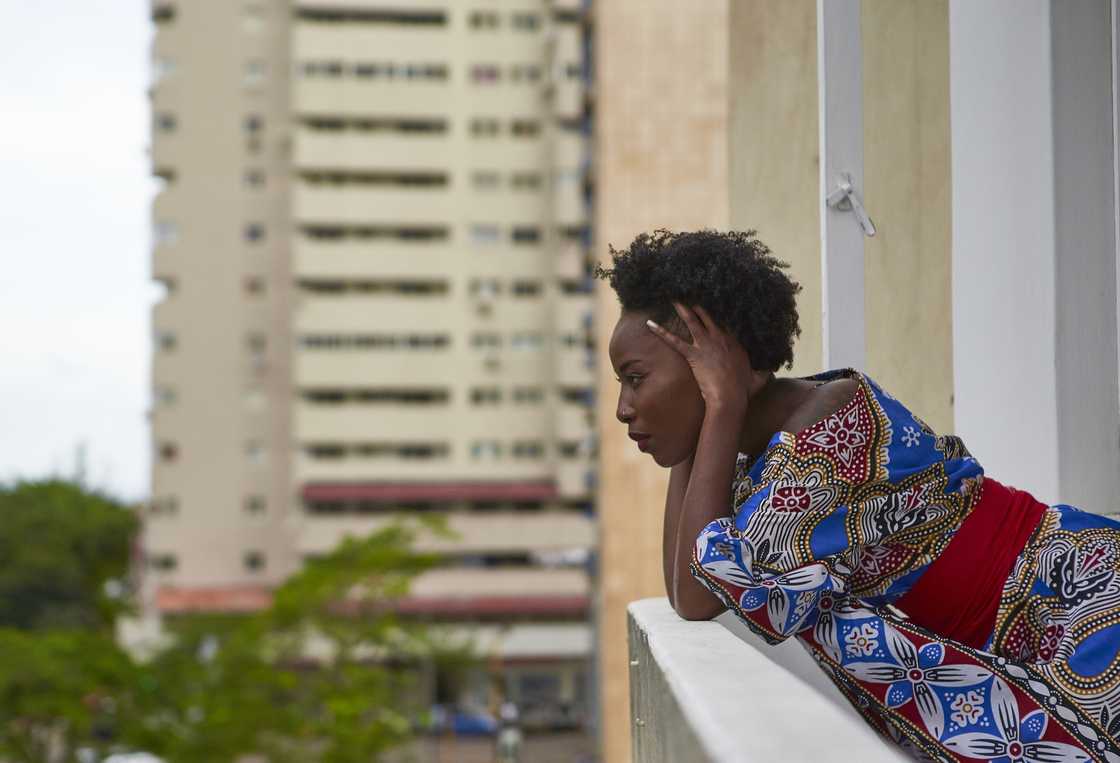Serious woman wearing colourful dress standing on balcony looking out.