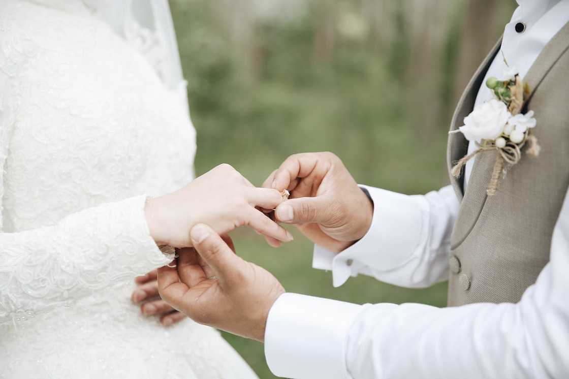 A close-up shows a groom placing a wedding ring on a bride’s finger during a ceremony.